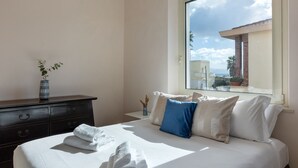 Un lit bien fait, avec du linge de lit blanc et un coussin bleu, une commode ornée d’un vase contenant des plantes séchées, et une fenêtre donnant sur des immeubles et des palmiers.