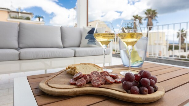 Une table en bois sur laquelle on trouve une planche à fromages, des tranches de salami et des raisins, deux verres de vin blanc, et un canapé gris en arrière-plan.