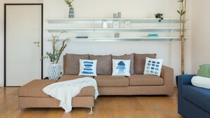 Un salon avec un canapé marron, un plaid blanc et un fauteuil bleu. Il y a des étagères au-dessus du canapé, où sont disposés des objets décoratifs, et une porte se trouve sur le côté gauche.