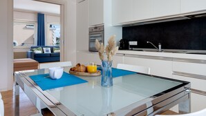 Une cuisine moderne avec des meubles blancs, une table à manger en verre et une vue sur un balcon agrémenté d’un canapé bleu.