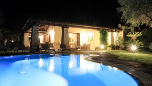 Una piscina con sillas de descanso y un patio, rodeada por un edificio bien iluminado de fachada de piedra y mobiliario exterior.