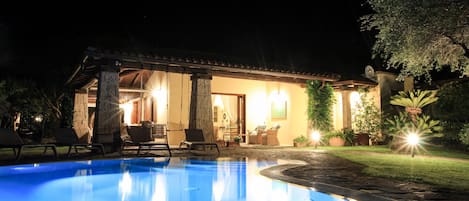 A poolside area with a clear pool, lounge chairs, and a building with warm lighting.