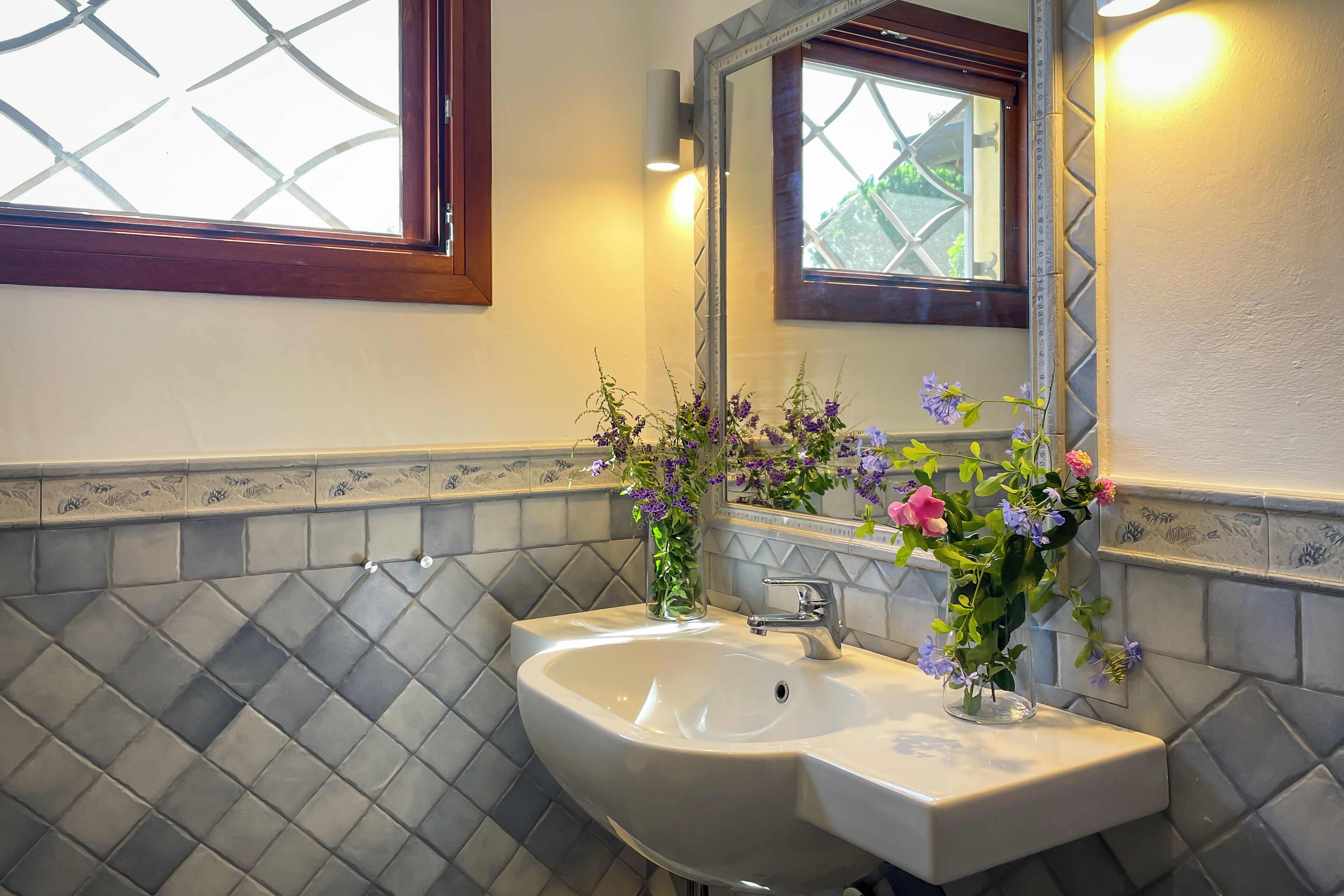 Un baño con un lavamanos blanco, dos floreros, un espejo y una ventana con marco de madera.