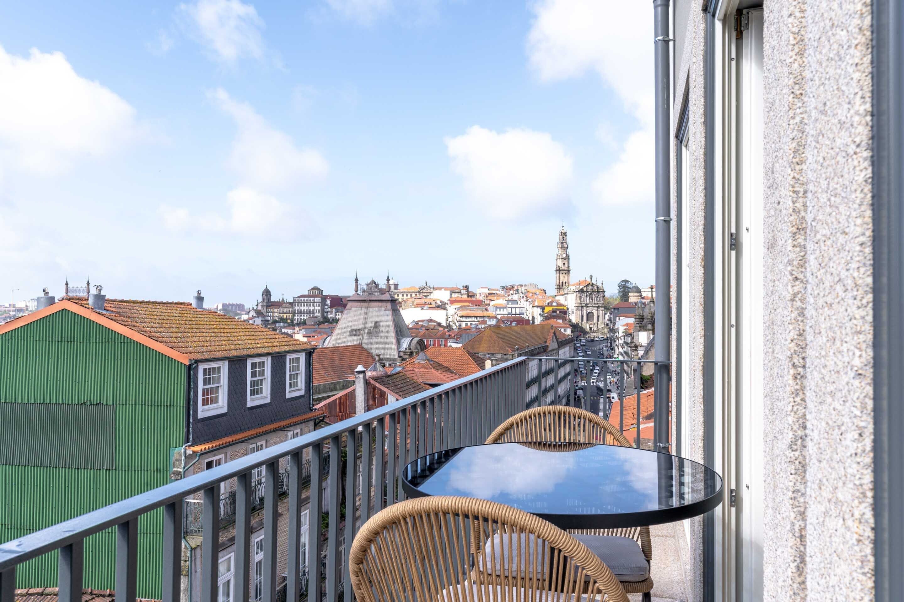 Un balcón con sillas de mimbre y una mesa de vidrio con vistas a un paisaje urbano que incluye edificios históricos y una torre destacada.