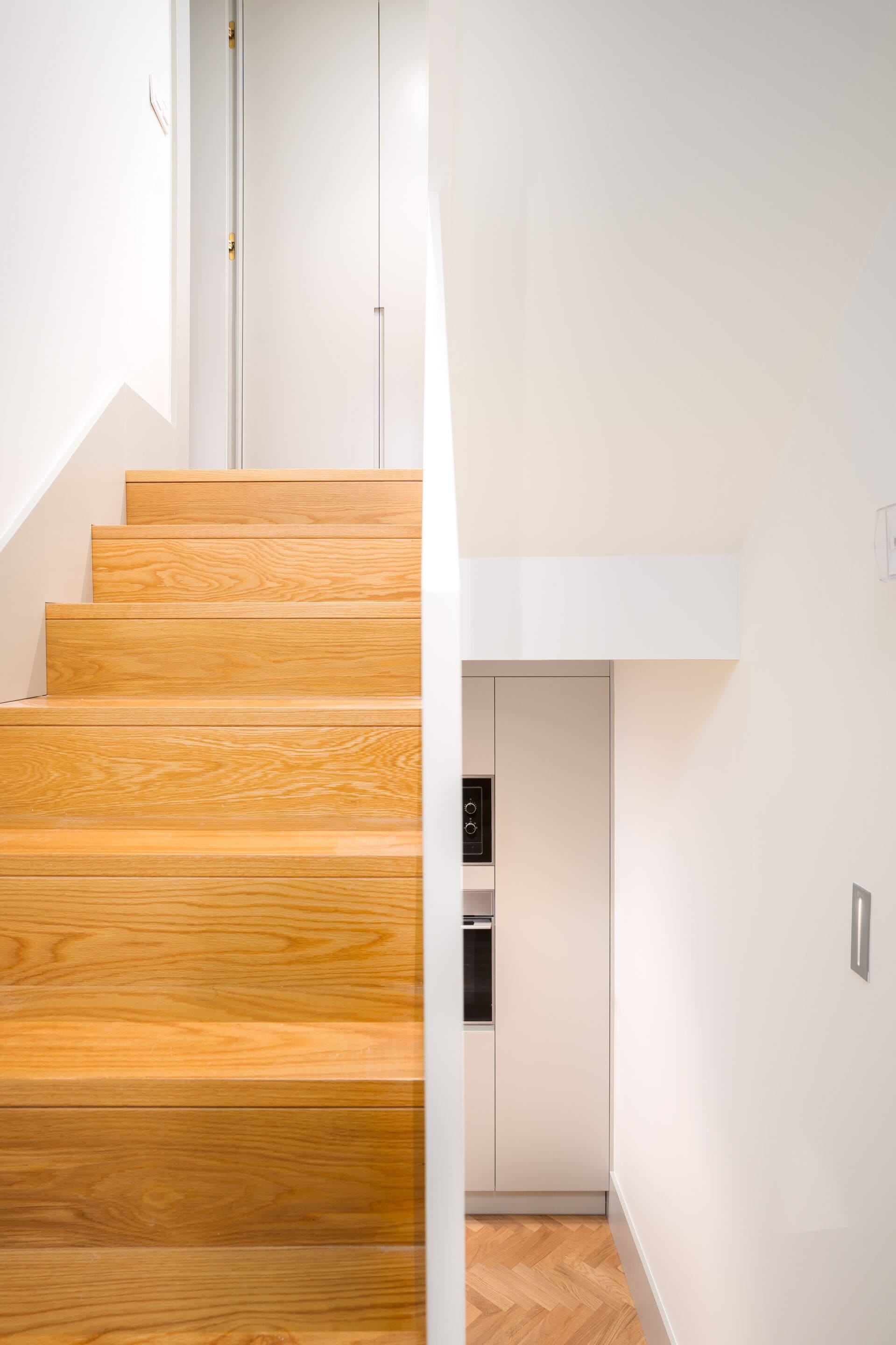 Un escalier en bois mène à une cuisine équipée de meubles blancs et d’un four encastré.