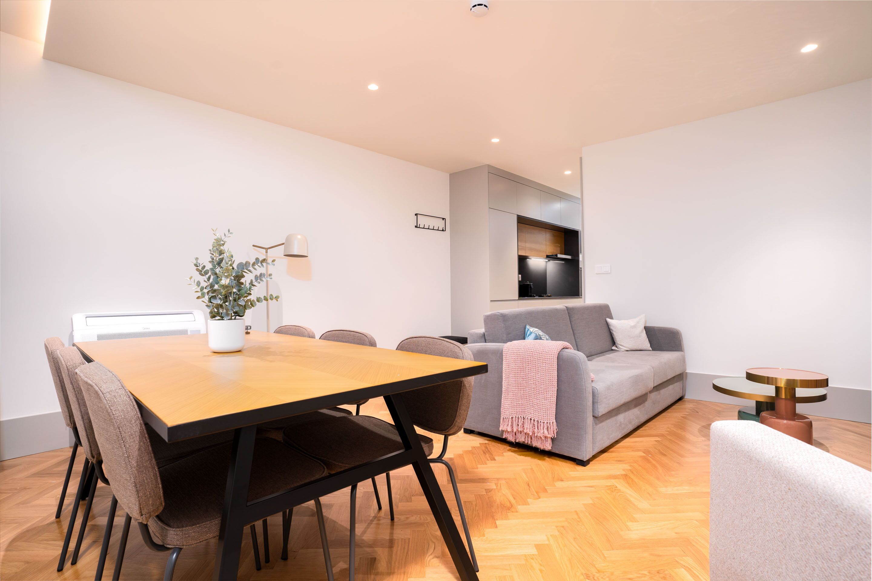 A modern living room with a wooden dining table, grey sofa, and a small round table with a lamp.