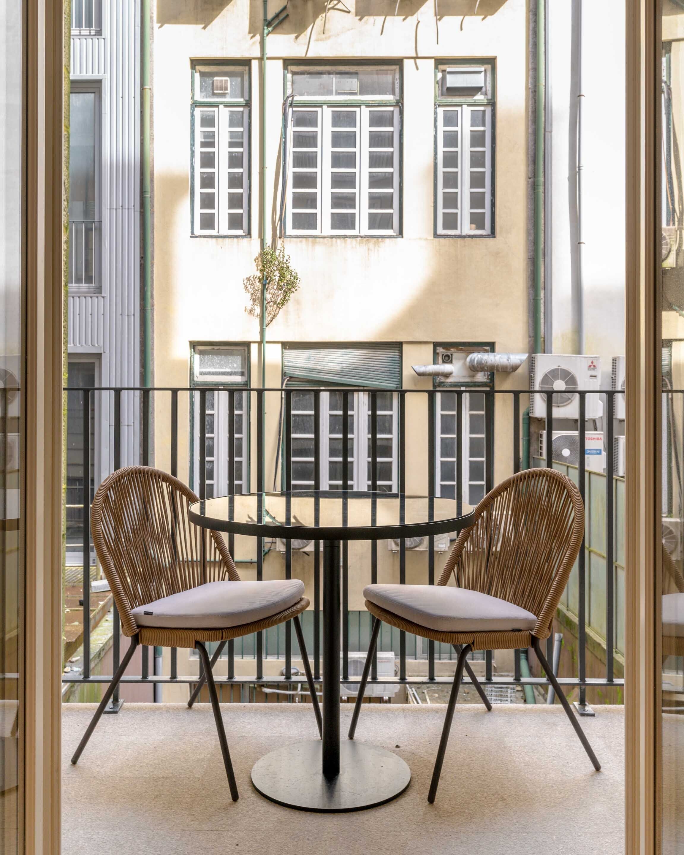 A balcony with a round table and two wicker chairs, overlooking a building with multiple windows and air conditioning units.