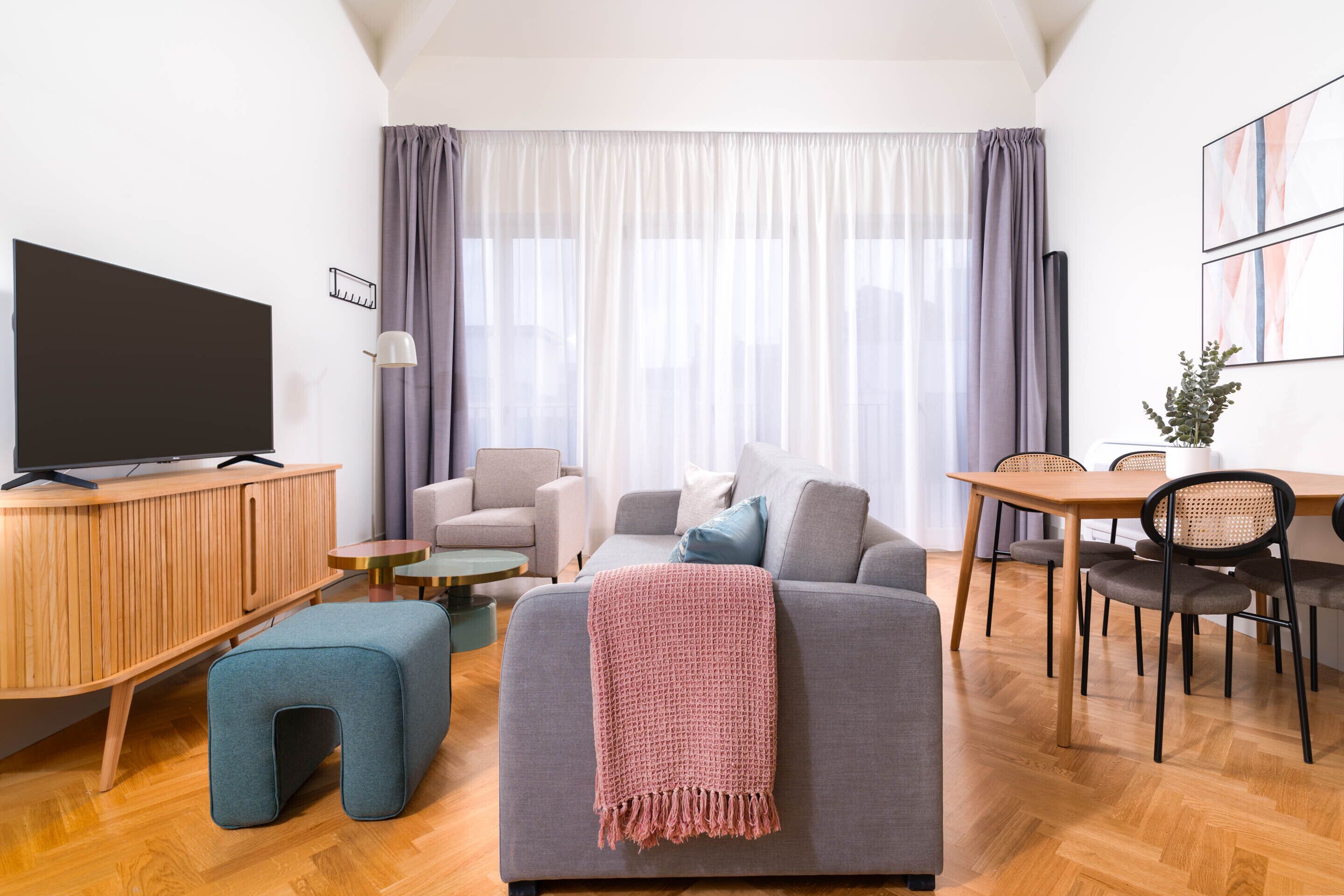 A modern living room with a grey sofa, a TV on a wooden cabinet, a small round table, and a dining area with a wooden table and chairs.