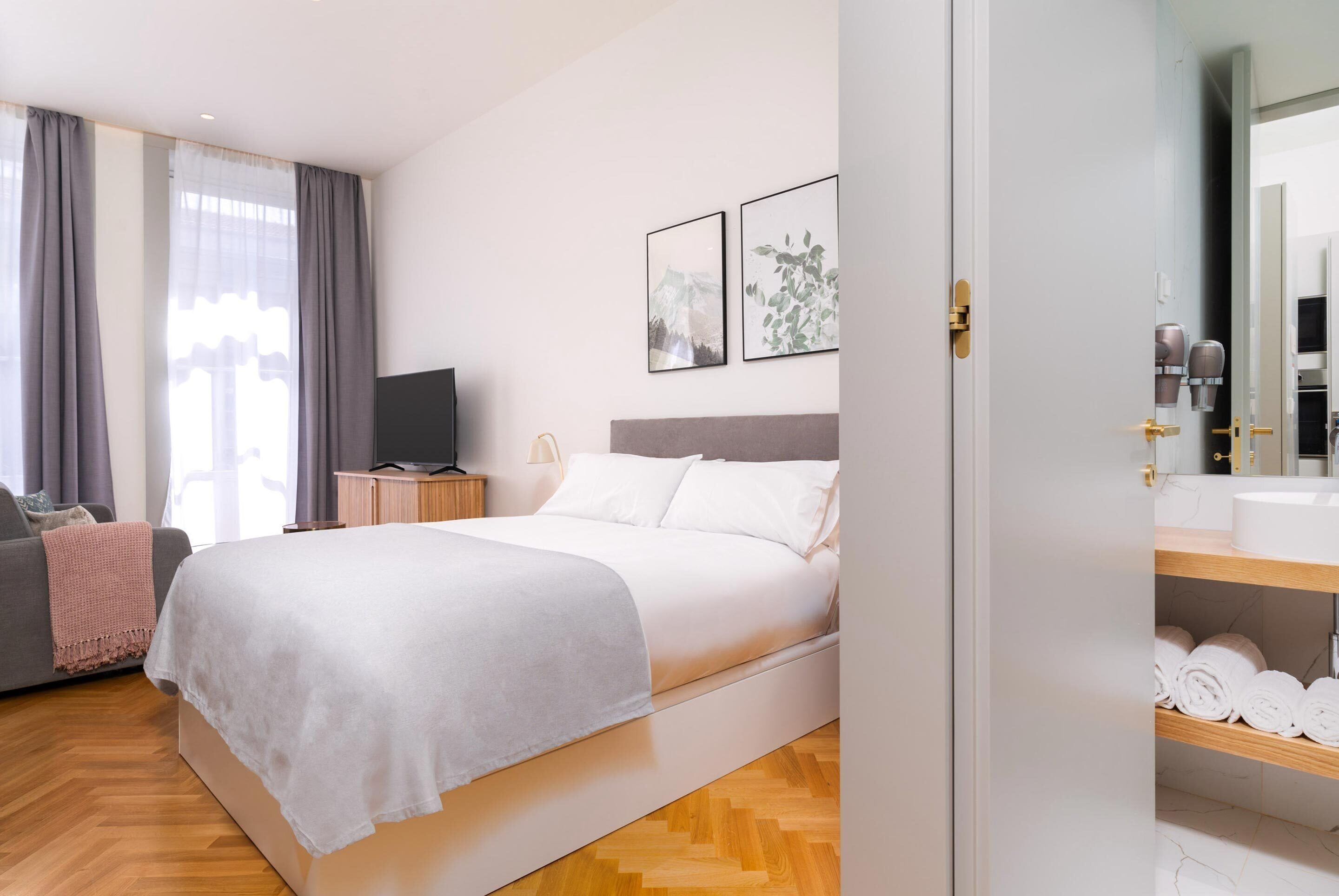 A modern bedroom with a bed, a TV, a bedside table, and a bathroom visible through an open door.