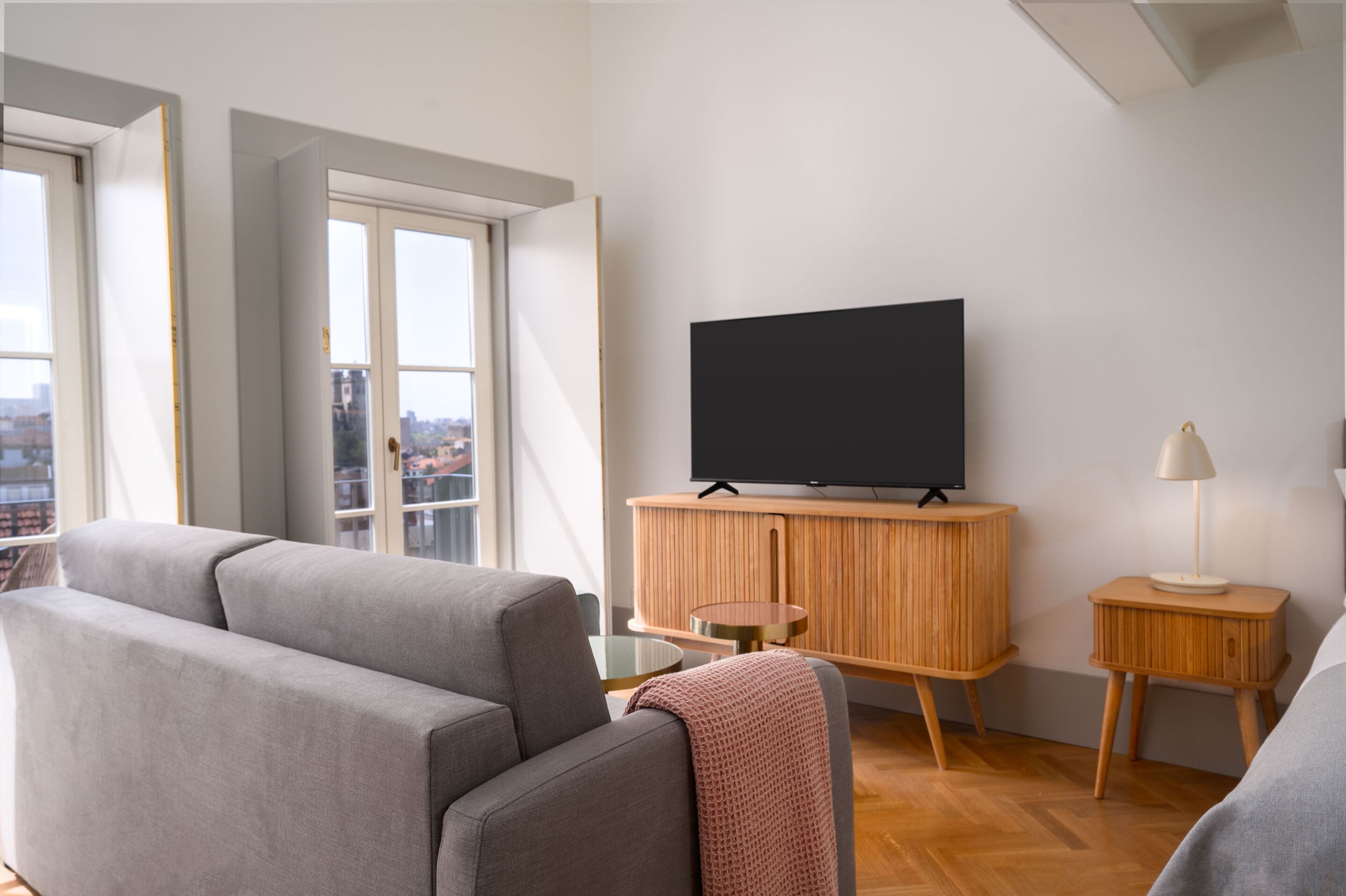 Uma sala de estar moderna com um sofá cinzento, um armário de madeira com uma TV, uma mesa redonda pequena e um abajur.