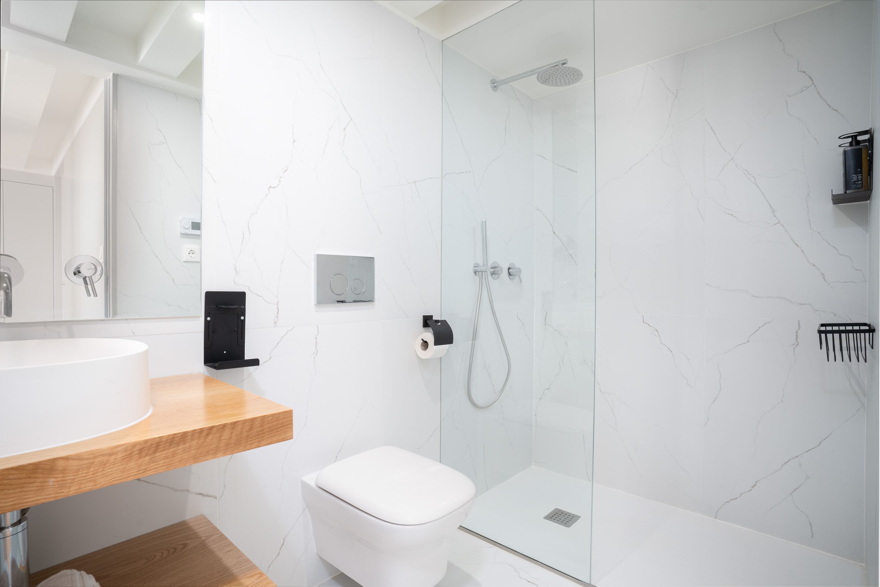 A modern bathroom with a white marble wall, a wooden vanity, a white toilet, and a glass-enclosed shower area.