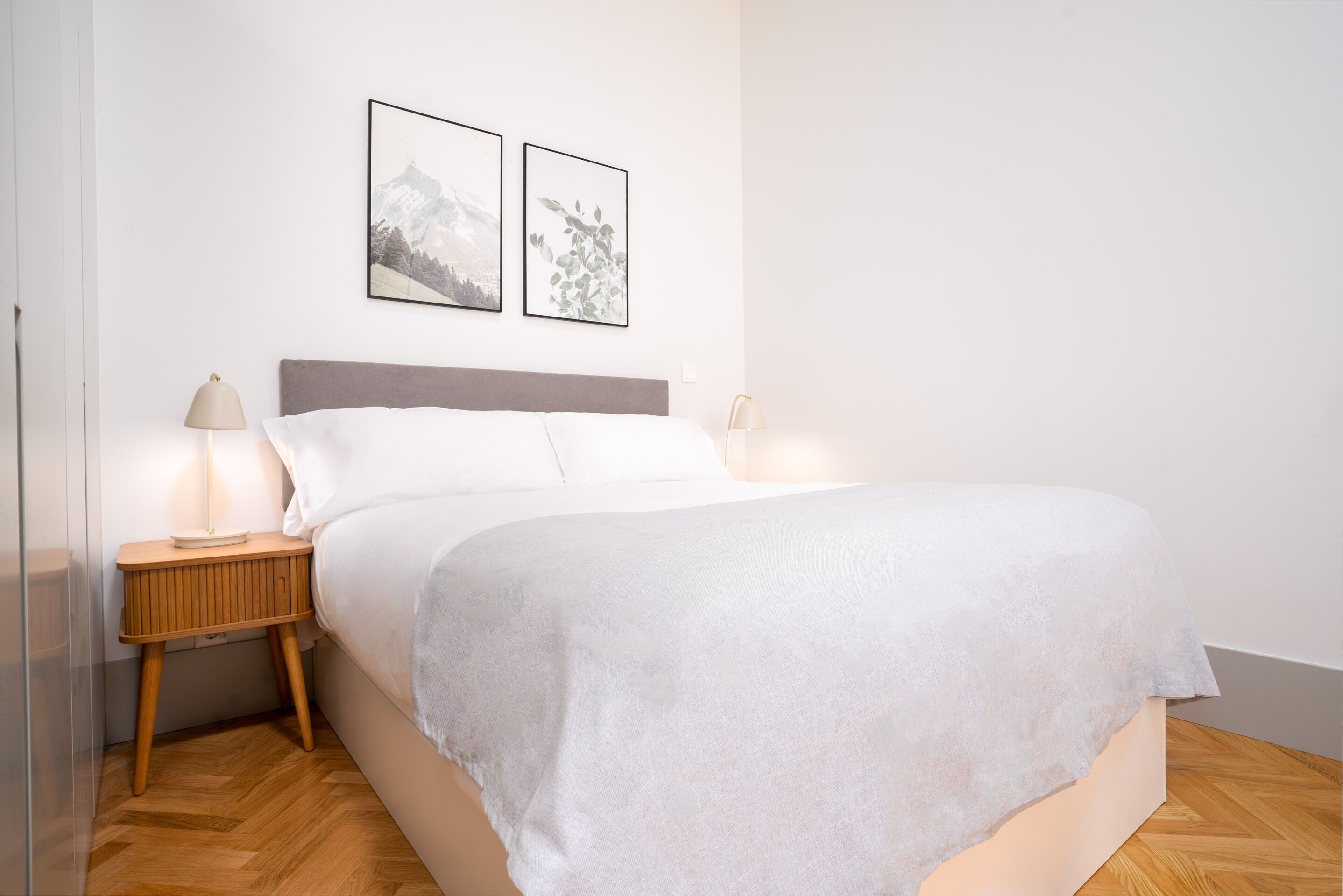 Una cama bien hecha con edredón blanco, una mesita de noche de madera con lámpara y dos cuadros en la pared.