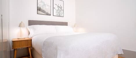 Una cama bien hecha con edredón blanco, una mesita de noche de madera con lámpara y dos cuadros en la pared.