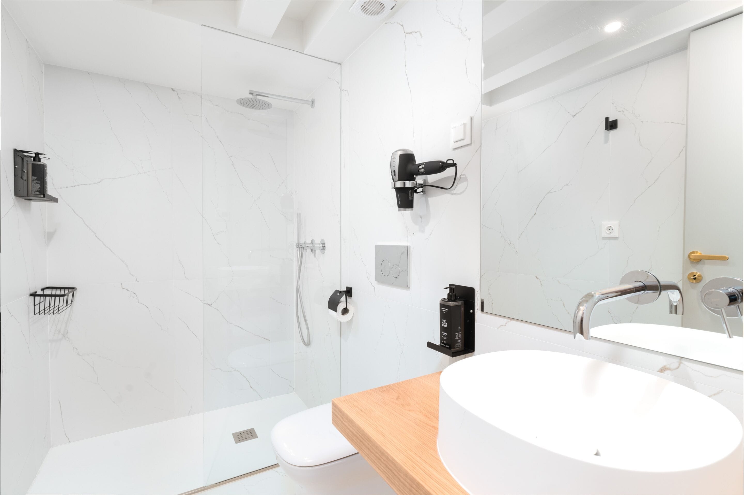 A modern bathroom with a white marble wall, a wooden vanity, a white sink, a handheld showerhead, and a towel rack.