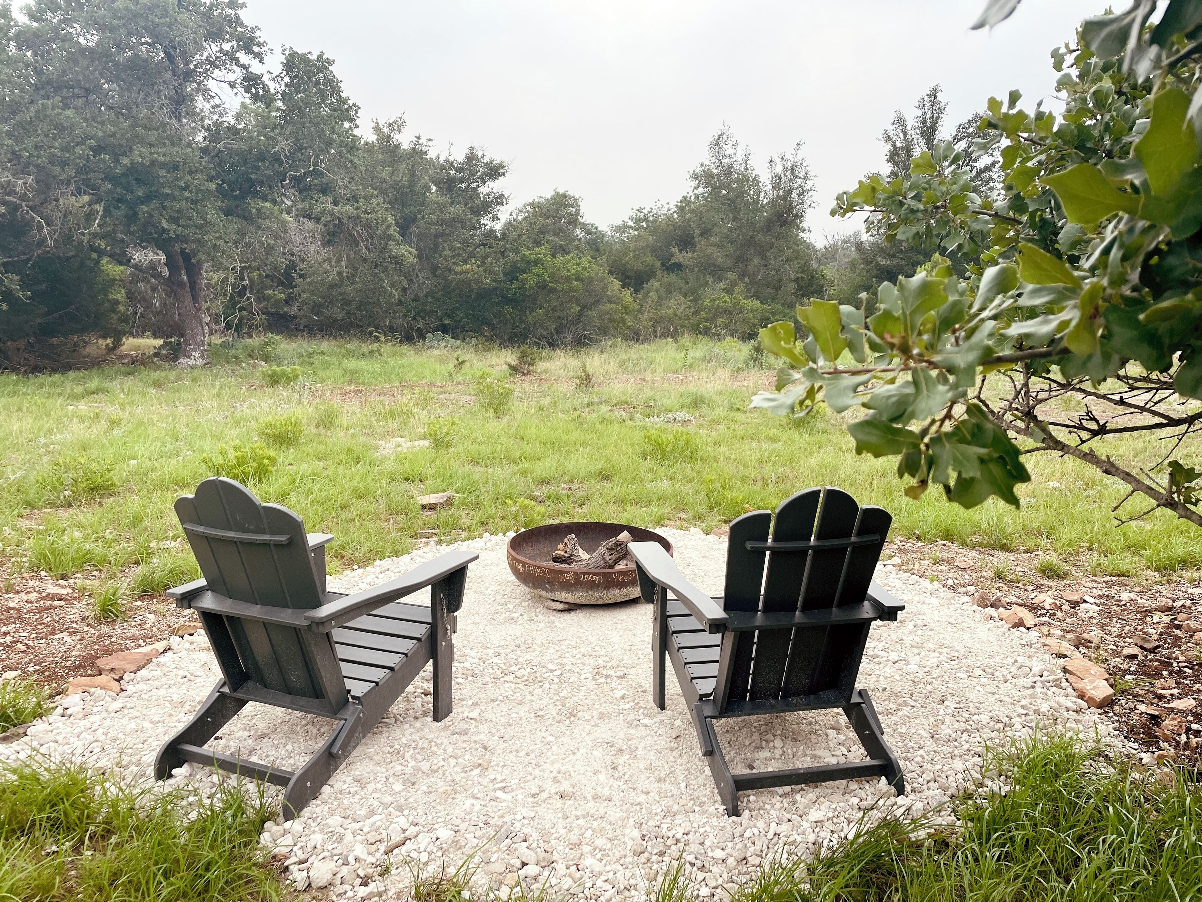 Deux fauteuils Adirondack sont placés face à un foyer, sur une surface recouverte de gravier, avec un champ herbeux et des arbres en arrière-plan.