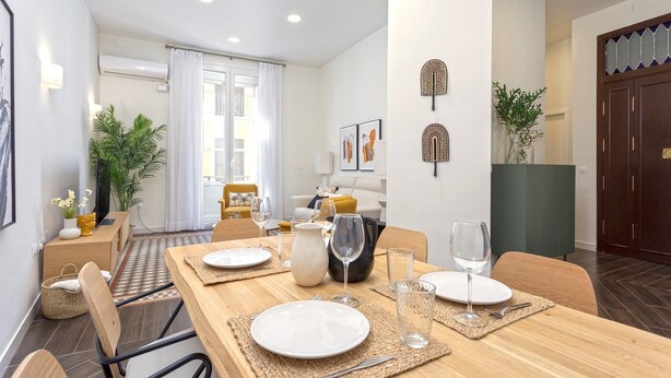 Un coin repas avec une table en bois dressée pour quatre personnes, entourée de chaises. On y trouve deux climatiseurs fixés au mur, une grande fenêtre avec des rideaux et un canapé à l’arrière-plan.
