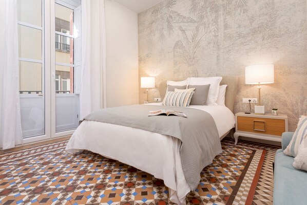 A bedroom with a bed, two bedside tables with lamps, a patterned floor, and a view through a window.