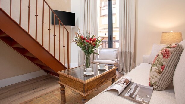 A living room with a wooden staircase, a glass coffee table with a vase of flowers, a sofa with a floral pillow, and a TV mounted on the wall.