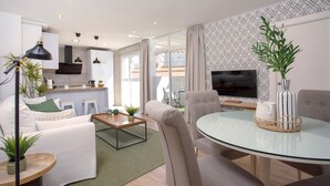Una sala de estar moderna con una mesa de vidrio, asientos cómodos, una cocina al fondo y un televisor montado en la pared.