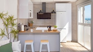 Una cocina moderna con una isla central, dos taburetes de bar, una lámpara colgante y vistas al mar a través de amplios ventanales.