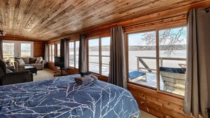 Habitación de cabaña de madera con una cama grande, un sofá, un televisor y vistas a un muelle y al agua.