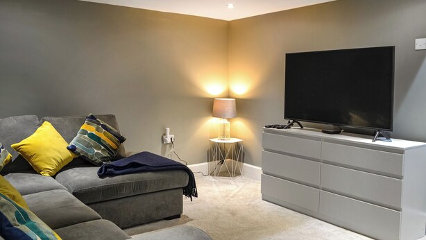 A modern living room with a grey sofa, a flat-screen TV, and a small side table with a lamp. - Doodles Holiday Home (Eyemouth)