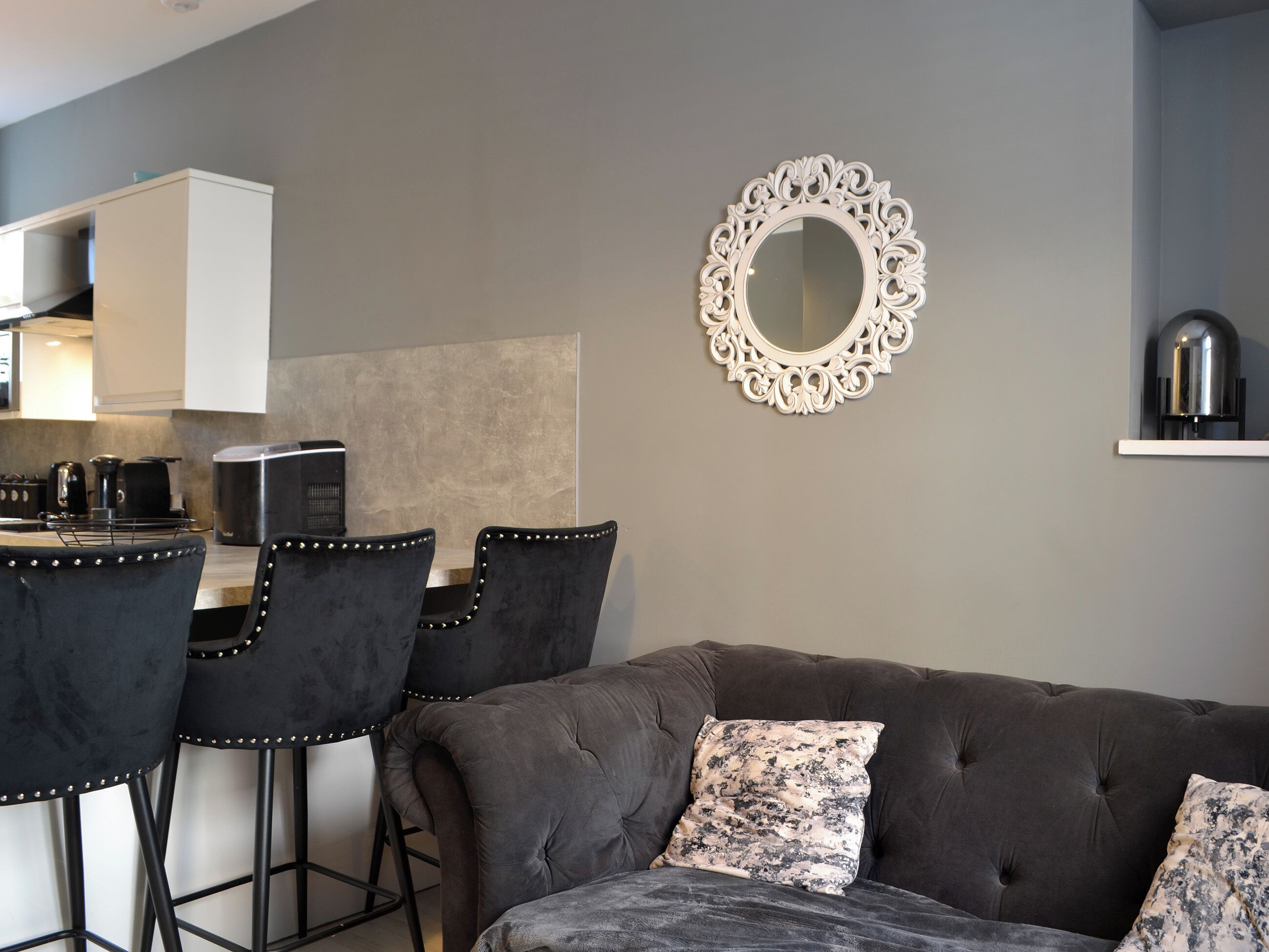 Un área de estar moderna con un sofá gris, una mesa de comedor con sillas y un espejo decorativo.