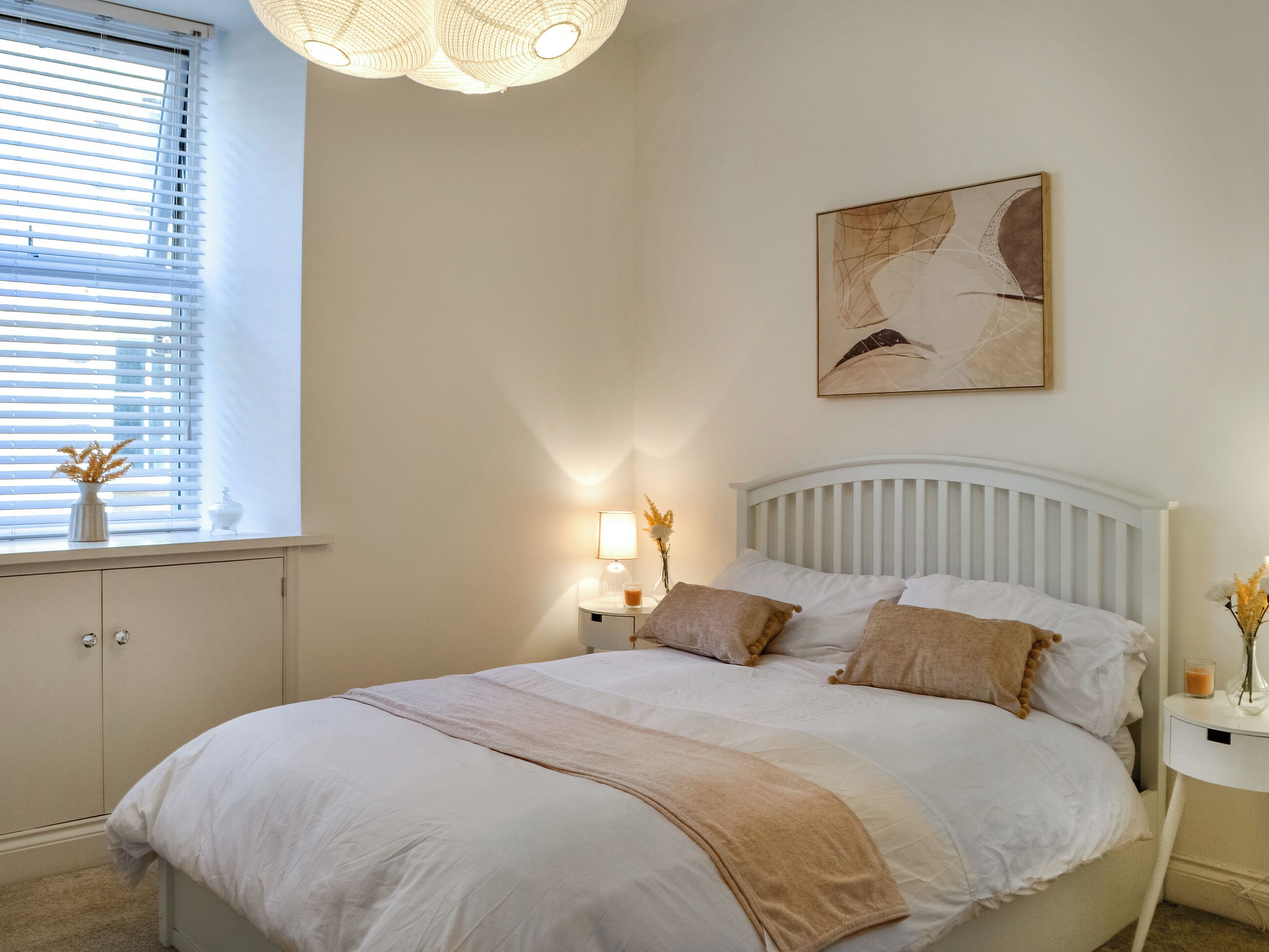Quarto com cama, mesas de cabeceira, janela com persianas, uma obra de arte na parede e luminária no teto.