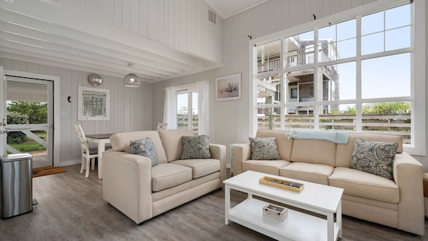 A spacious living room with two beige sofas, a white coffee table, and a dining area with a table and chairs. Large windows offer views of a building under construction. - Oceanside Cottage Steps from the Beach! The Gray Sandcastle! (Nags Head)