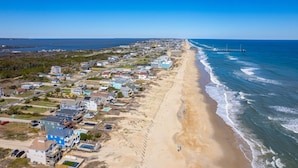 An aerial view of a coastal town with beachfront properties, a pier extending into the ocean, and a clear blue sky. - Oceanside Cottage Steps from the Beach! The Gray Sandcastle! (Nags Head)