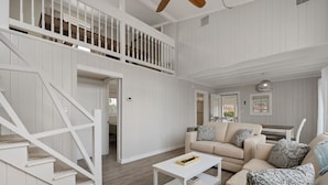 A spacious living area with a high ceiling, white wooden walls, a beige sofa set, a white coffee table, and a staircase with white railings. - Oceanside Cottage Steps from the Beach! The Gray Sandcastle! (Nags Head)