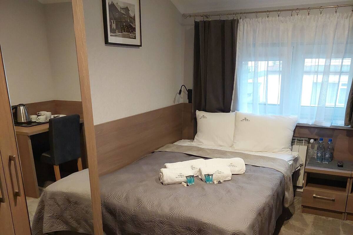 A neatly arranged hotel room with a bed, a desk, and a chair. There is a window with curtains, a framed picture on the wall, and a small table with a kettle and cups.