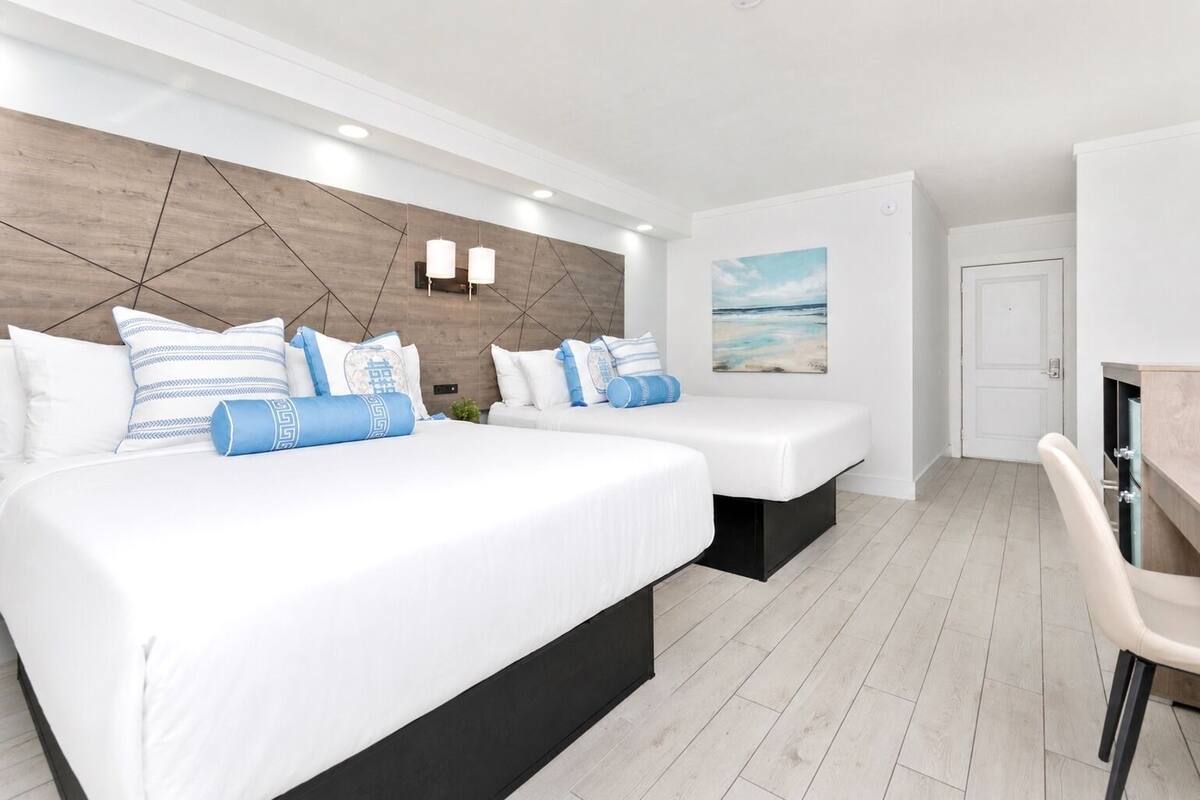 Habitación de hotel con dos camas, un escritorio, una silla, un cuadro en la pared y un cabecero de madera.