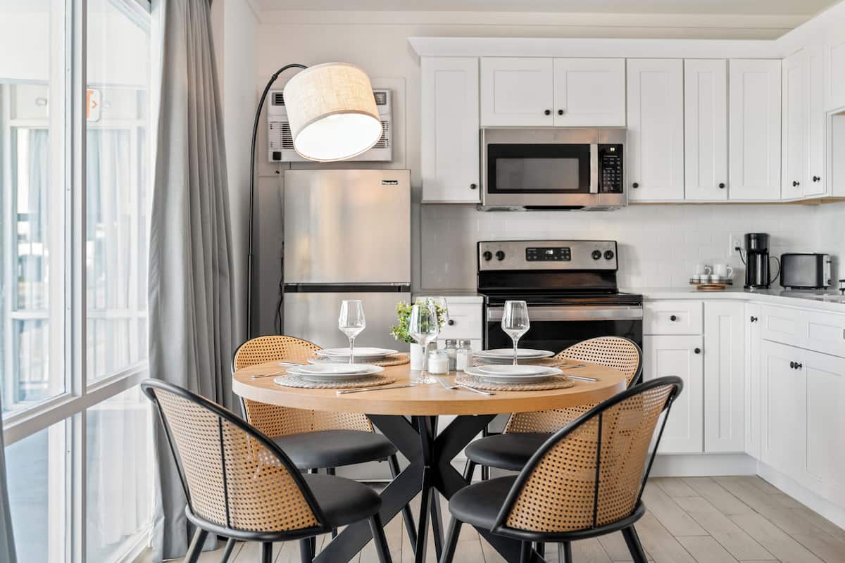 Una cocina moderna con armarios blancos, un refrigerador de acero inoxidable, un microondas y un horno. Hay una mesa de comedor con sillas y una lámpara encima.