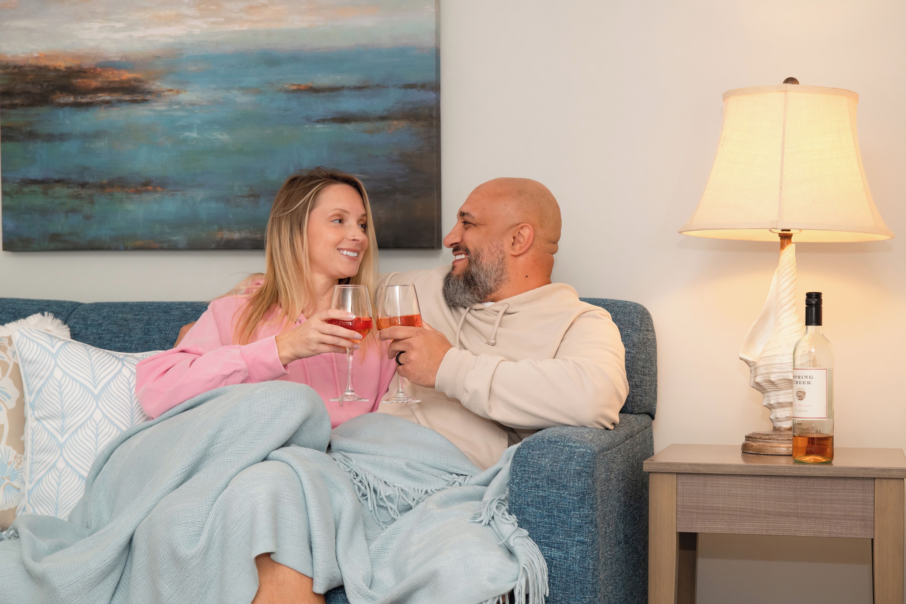 A couple sitting on a couch with a painting in the background, a lamp, and a bottle on a side table.
