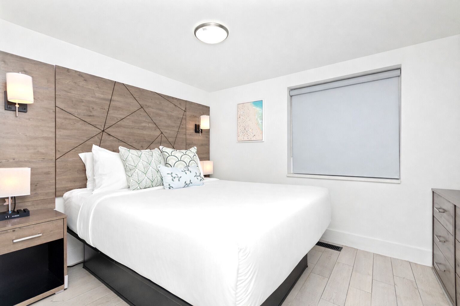 A modern bedroom with a large bed, bedside tables, a headboard with geometric patterns, wall-mounted lights, and a window with blinds.