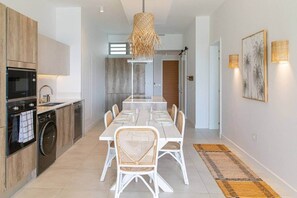 Une cuisine moderne dotée d’appareils électroménagers intégrés, un coin repas avec des meubles blancs et un grand luminaire tressé.