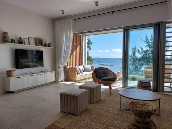 Un salon moderne avec un téléviseur, un canapé et un fauteuil en osier, de grandes fenêtres offrant une vue sur la mer, et un balcon aménagé avec du mobilier d’extérieur.