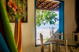 Quarto com vista para a praia através de uma janela, uma cadeira de madeira e um quadro na parede.