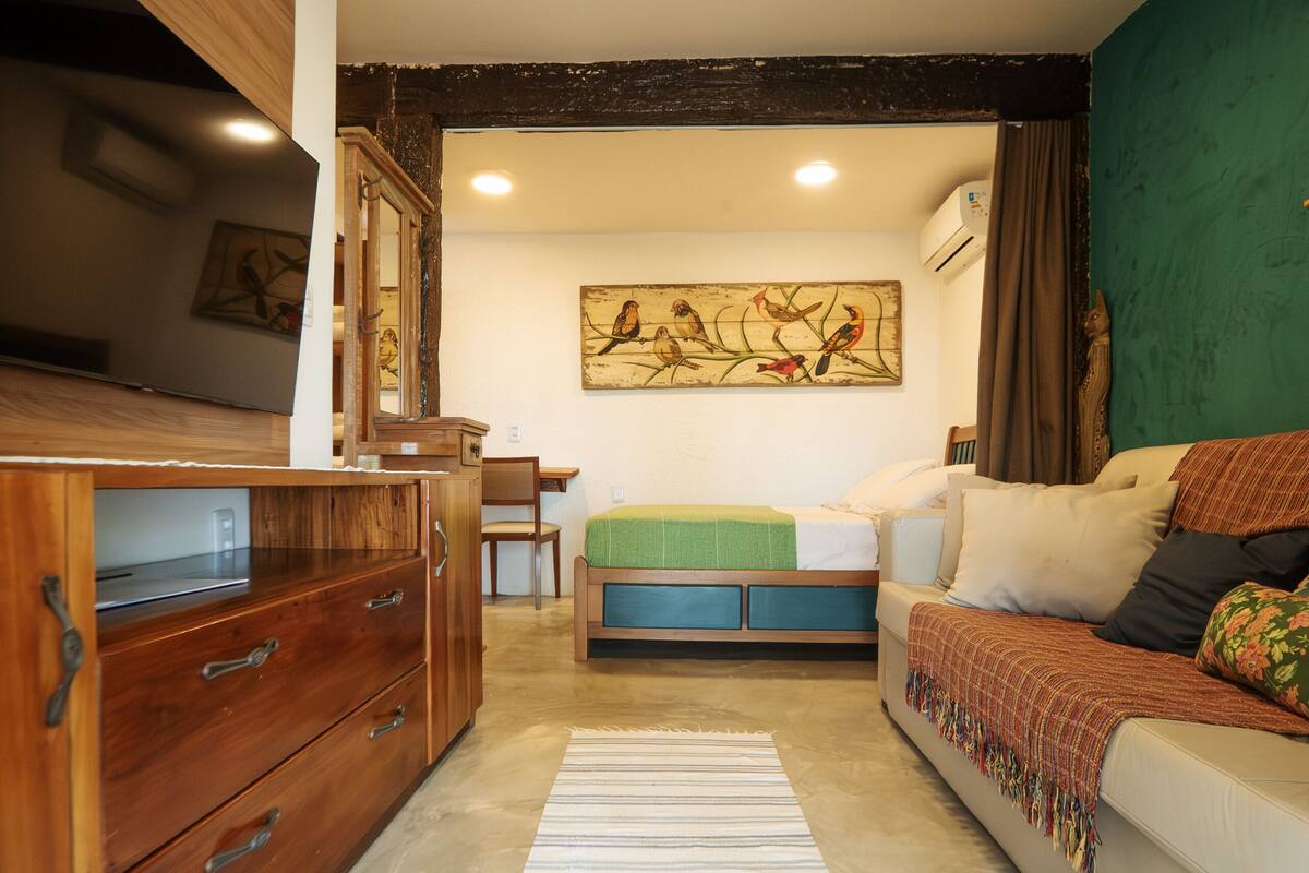 Une pièce chaleureuse avec une commode en bois, un lit recouvert d’une couverture verte, un tableau au mur et un luminaire au plafond.