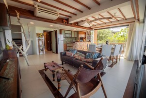 Uma sala de estar ampla com sofá, cadeiras, uma mesa de jantar e uma cozinha ao fundo.