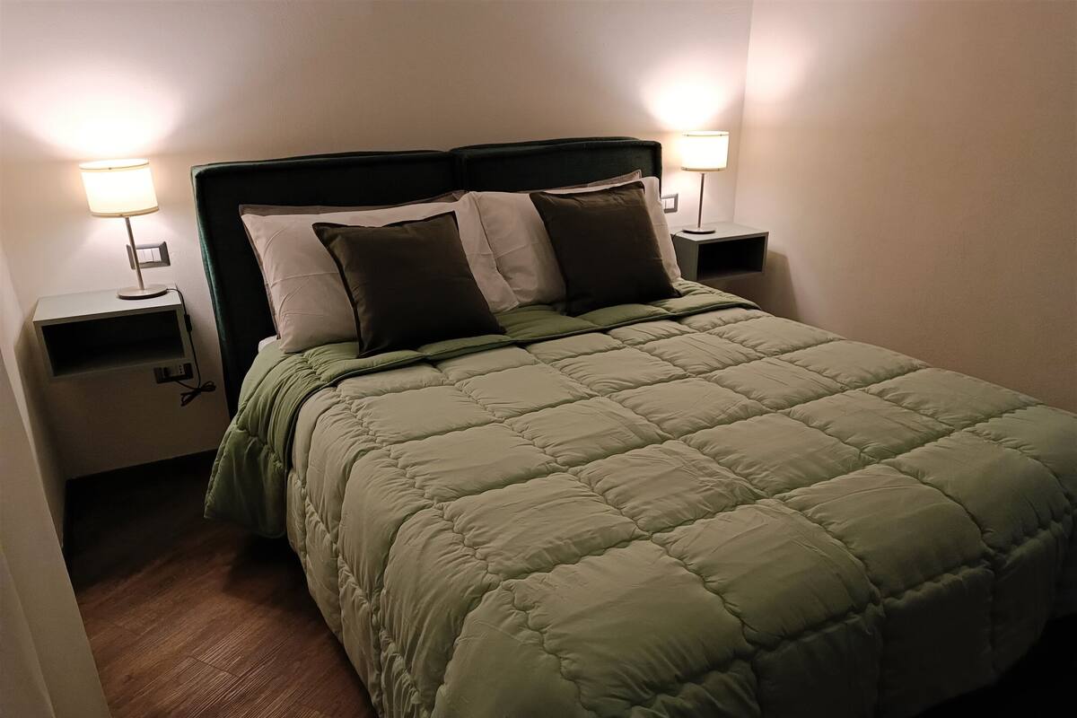 Una cama bien hecha con un edredón verde, dos lámparas de noche y suelo de madera.