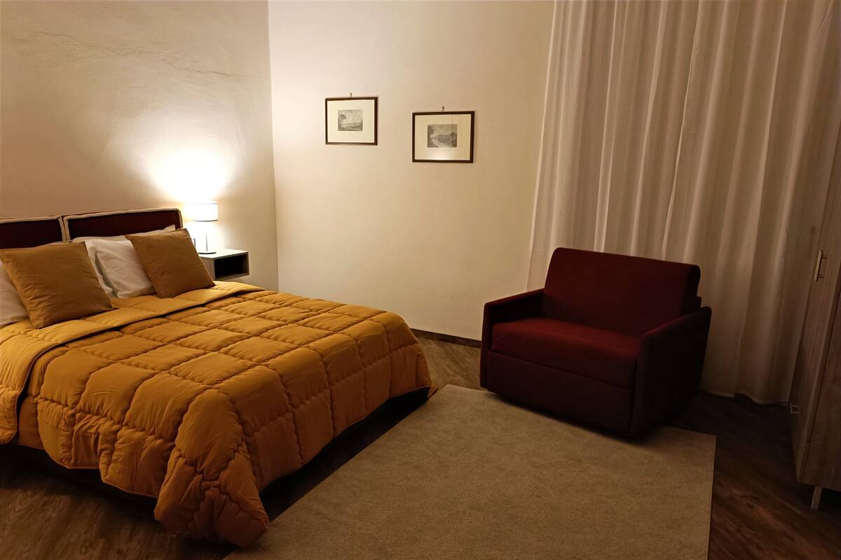 Una habitación de hotel con una cama, un sillón rojo, una mesita de noche y dos cuadros en la pared.