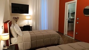 Quarto de hotel com duas camas, uma televisão presa à parede, uma escrivaninha com abajur e um banheiro visível por uma porta aberta.