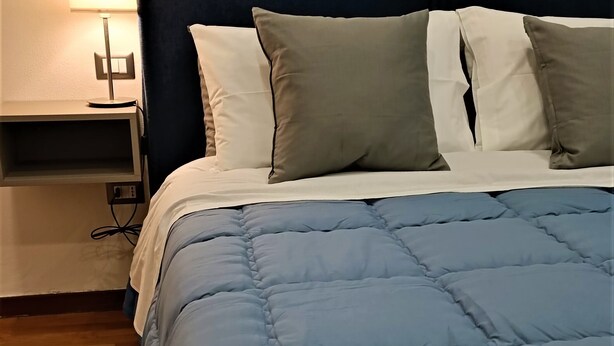 Uma cama bem arrumada com um edredom azul, uma luminária de cabeceira e uma mesa de cabeceira com abajur.