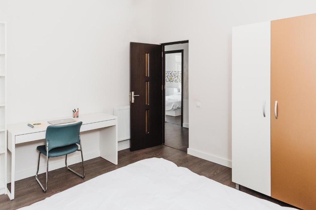 A bedroom with a white bed, a desk with a chair, a wardrobe, and a door leading to a bathroom.