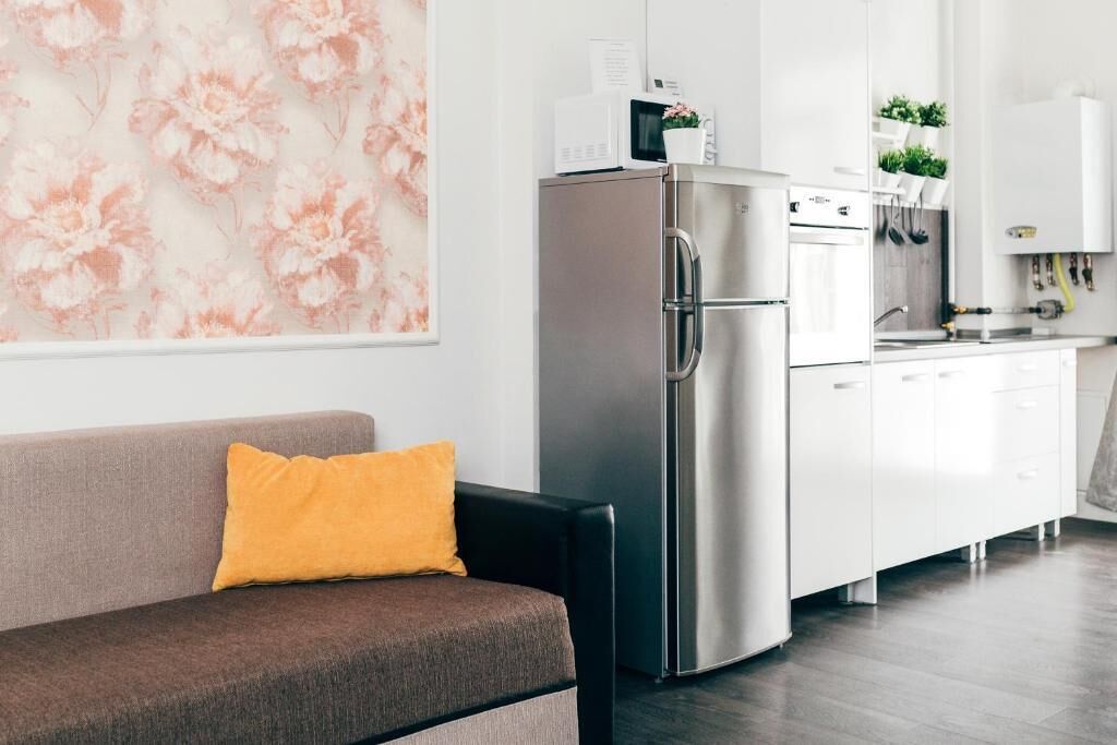 A modern kitchen with a stainless steel refrigerator, white cabinets, and a grey sofa with a yellow pillow.