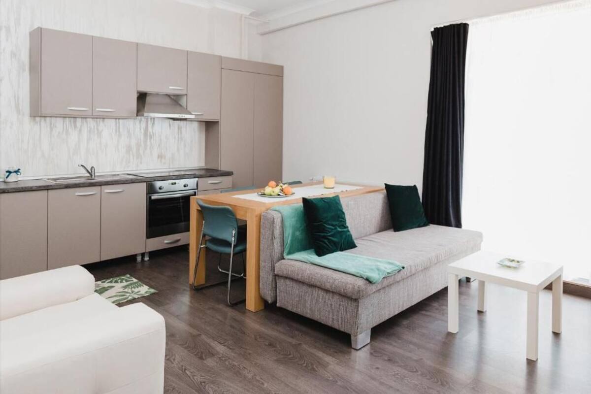 A modern living room with a grey sofa, a wooden dining table, and a kitchen area with cabinets and appliances.