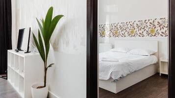 Un dormitorio moderno con una cama blanca, una mesita de noche, una planta en maceta y una consola blanca con un televisor.