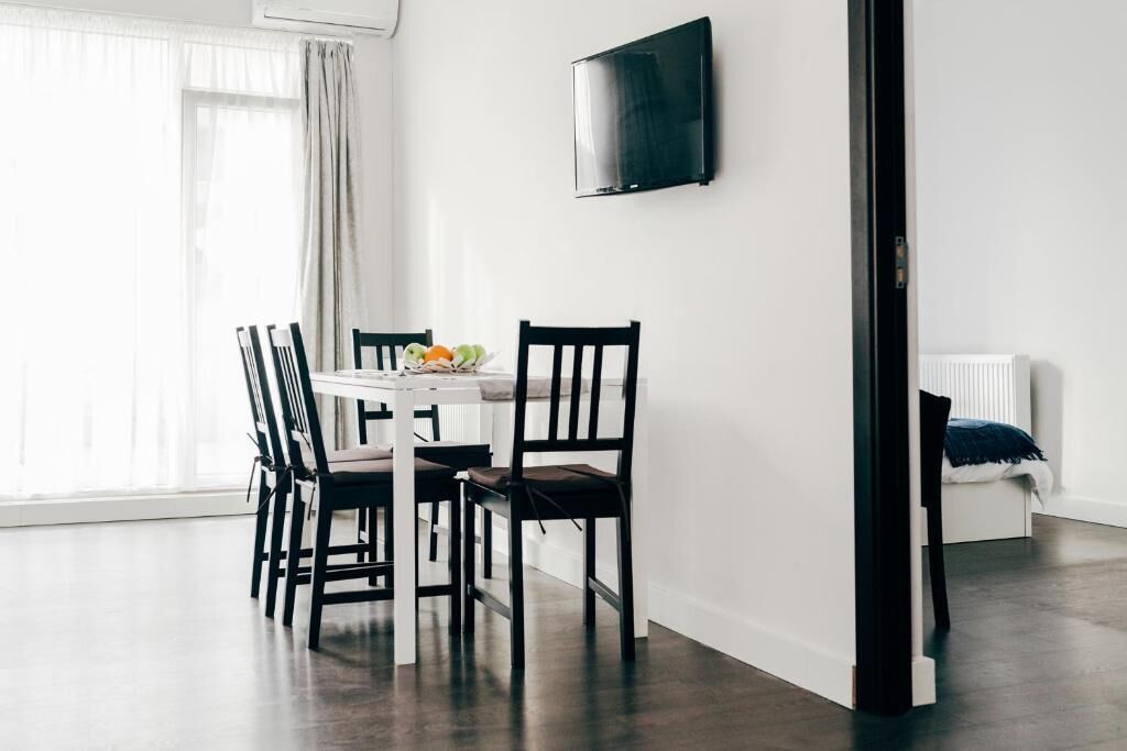 A modern dining area with a white table and black chairs, a flat-screen TV on the wall, and a window with sheer curtains.